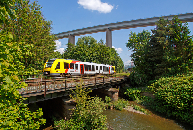 Un treno HLB bianco e giallo attraversa un ponte che porta sopra un fiume in una foresta.