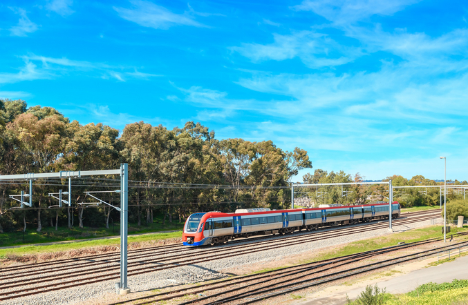 Un treno Keolis rosso e argento si muove da destra a sinistra davanti ad alberi e a un cielo blu brillante.